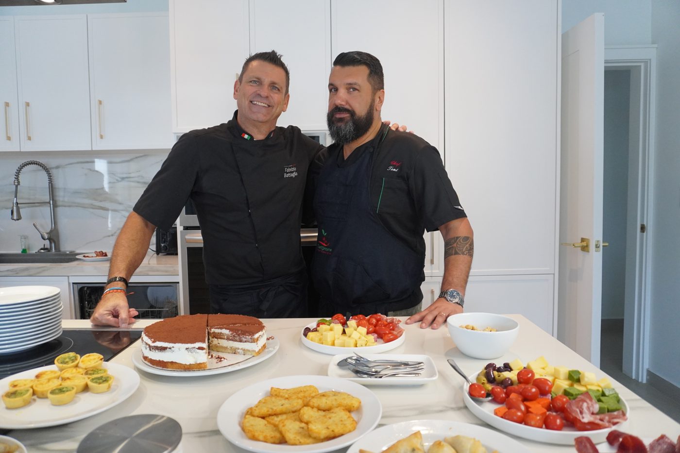 Fabrizio and Tony side by side in a bright kitchen, with a tiramisu and an antipasto spread on the counter