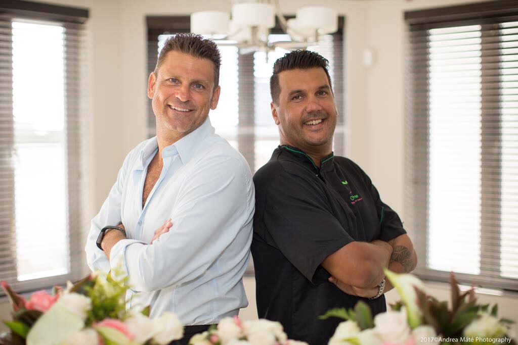 Two chefs smiling, standing back to back in a bright dining room