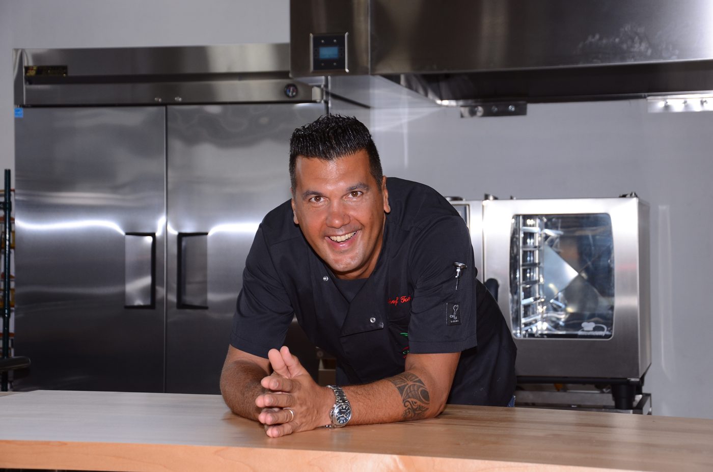 Chef Tony smiling, leaning on a wooden counter in a professional kitchen